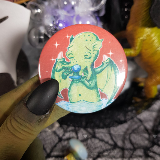 Hand holding a 58mm Cthulhu badge with a red background and sparkles, featuring Cthulhu drinking tea. Ideal for HP Lovecraft fans.