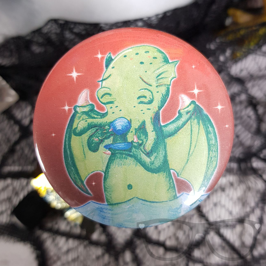 Close-up of a 58mm Cthulhu badge with a red background and sparkles, featuring Cthulhu drinking tea. Perfect for HP Lovecraft fans.