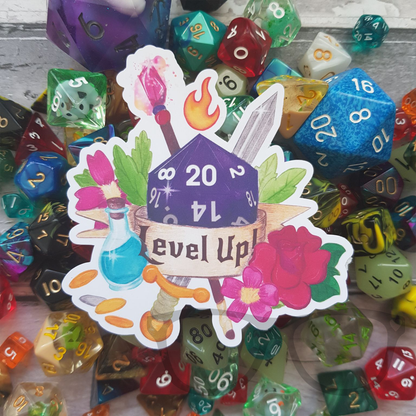 Level up with this glossy vinyl sticker. A purple dice surrounded by items you'd find when playing tabletop fantasy RPGs and a ribbon with the words "Level Up!" written on it.