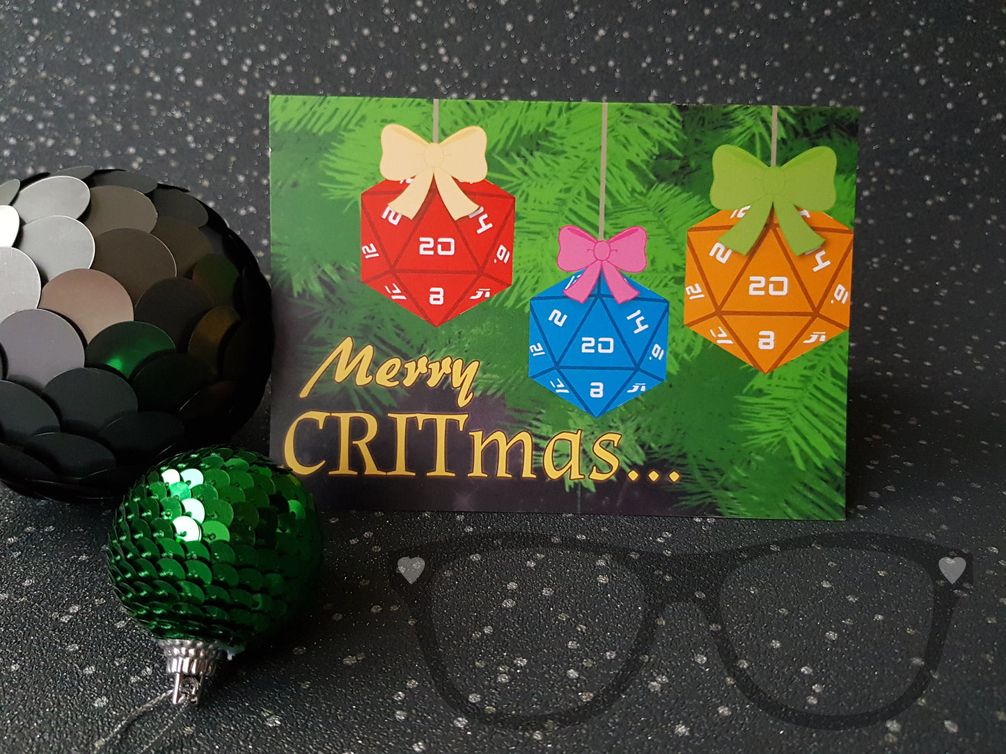 A Christmas greeting card that shows a red, blue and yellow 20 sided dice hanging from a festive tree. The text Reads "Merry Critmas" 