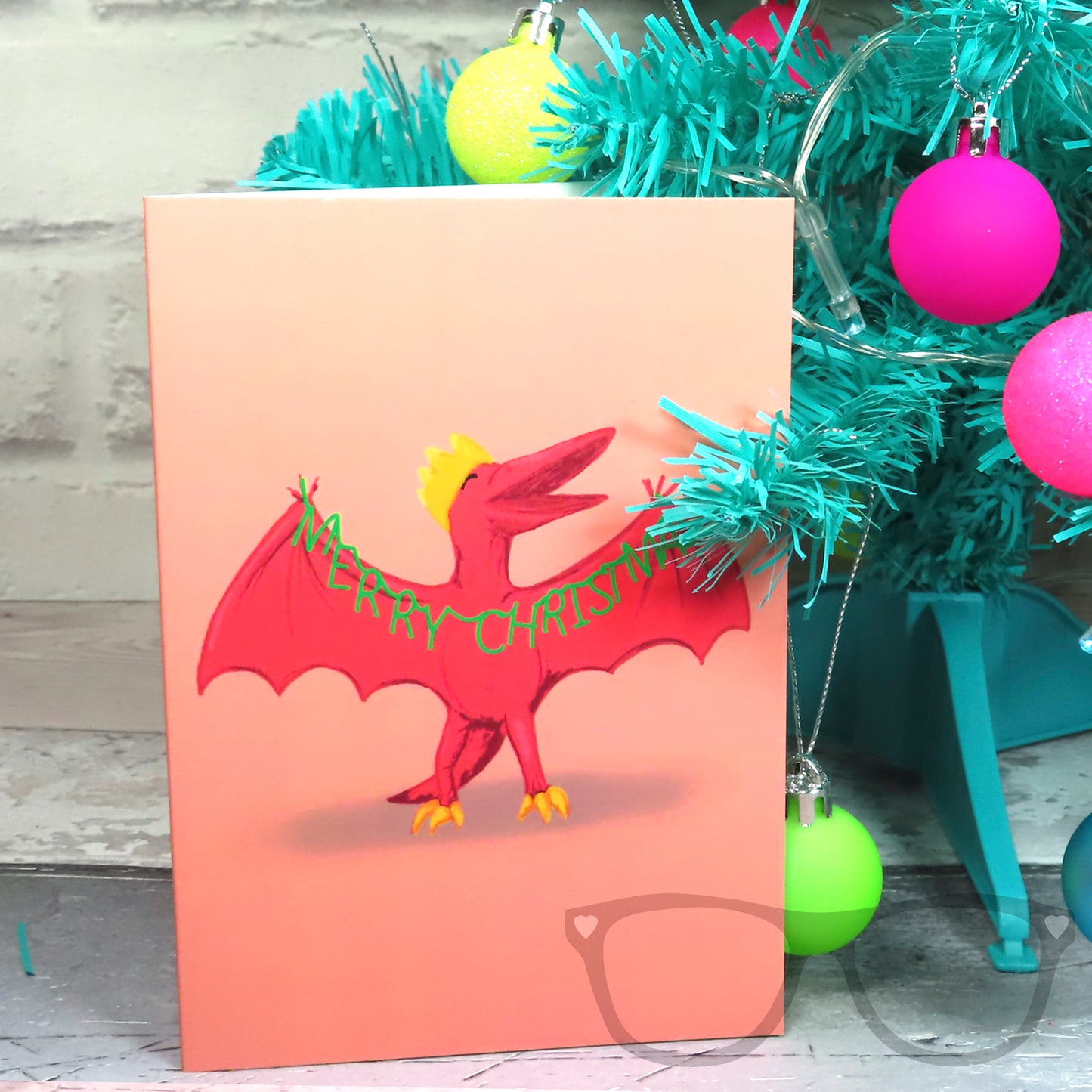 Dinosaur Christmas Card stood against a Christmas tree, features Pterry the Pteranodon holding a green Merry Christmas Garland 