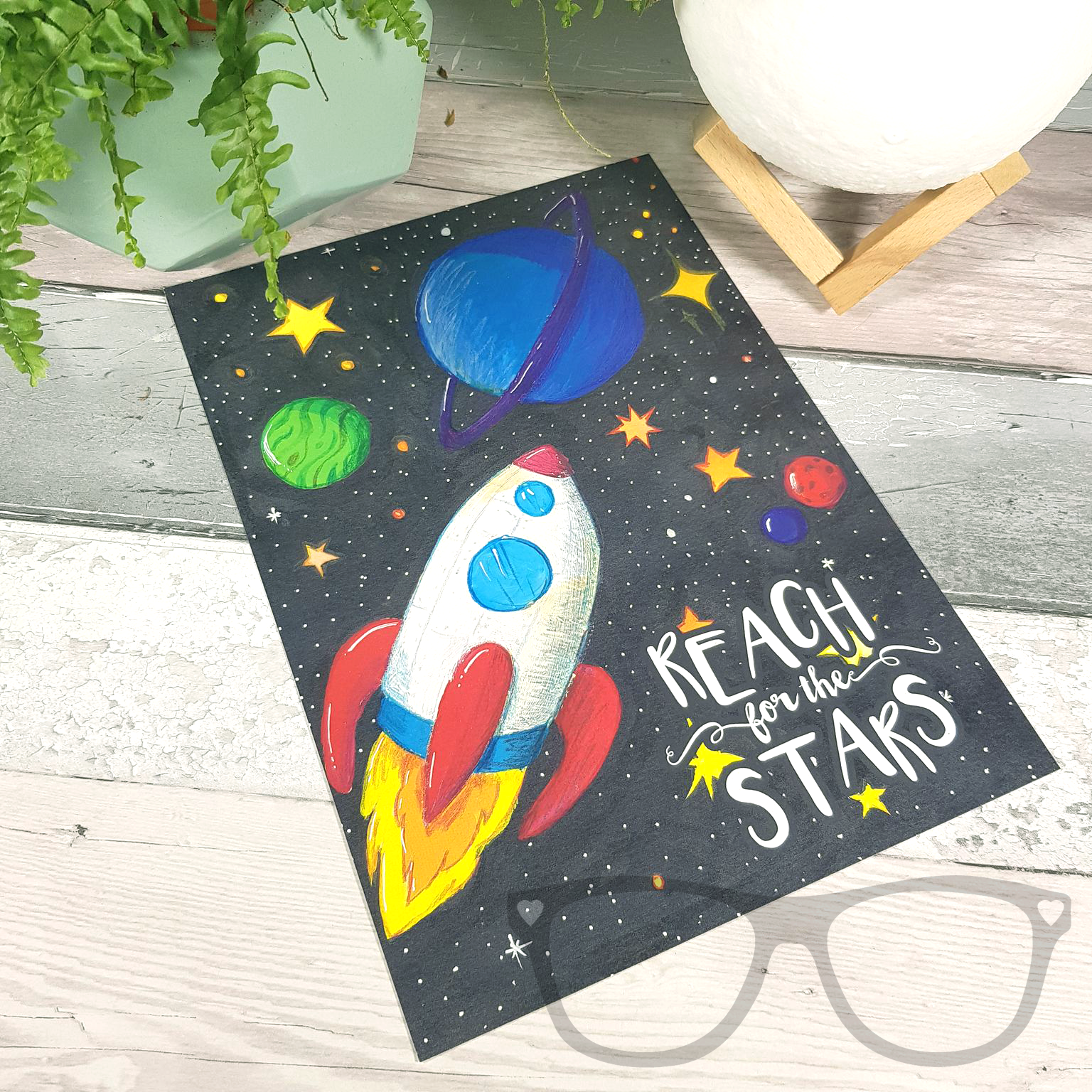 Reach for the stars A4 art print, a digital print of an original illustration showing a white rocket shooting through space with p[lanets and stars. The text reads "Reach for the stars"