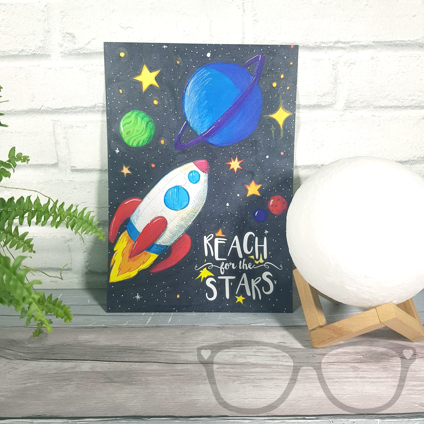 Reach for the stars A4 art print, a digital print of an original illustration showing a white rocket shooting through space with p[lanets and stars. The text reads "Reach for the stars" it is shown without a frame 