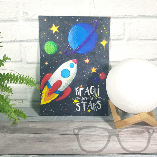 Reach for the stars A4 art print, a digital print of an original illustration showing a white rocket shooting through space with p[lanets and stars. The text reads "Reach for the stars" it is shown without a frame 