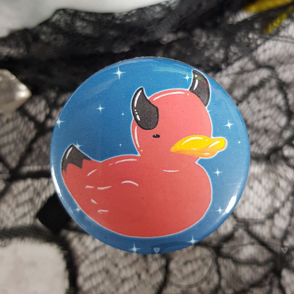 The 58mm circular badge shows a red duck with black devil horns on a teal background surrounded by little sparkles 
