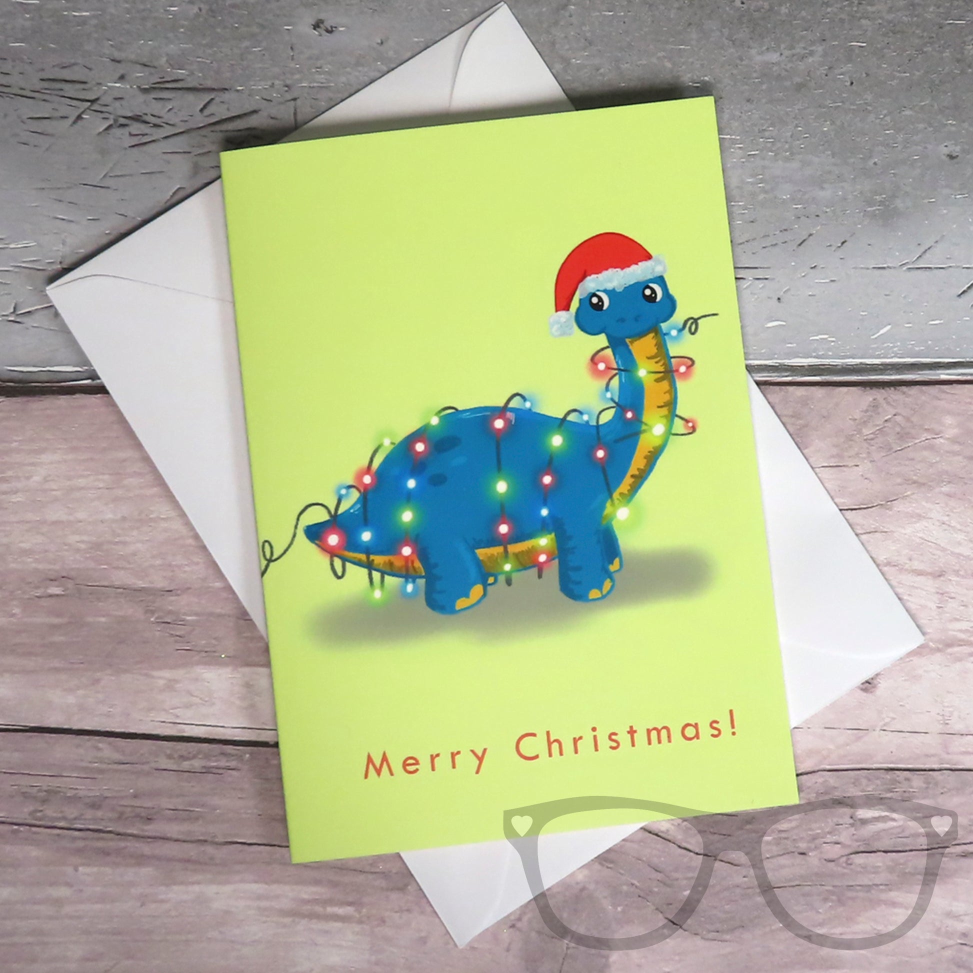 Christmas Card with a dinosaur tangled in fairy lights, a green brachiosaurus is standing on a yellow background wearing a festive santa hat. The card lays flat with the white envelope underneath
