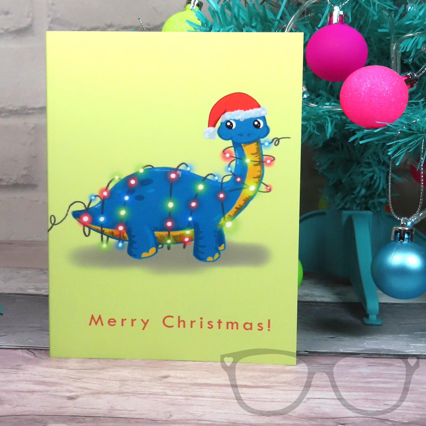 Brenda the Brachiousaurs is tangled up in fairy lights wearing a festive hat. A cute design on a christmas greetings card