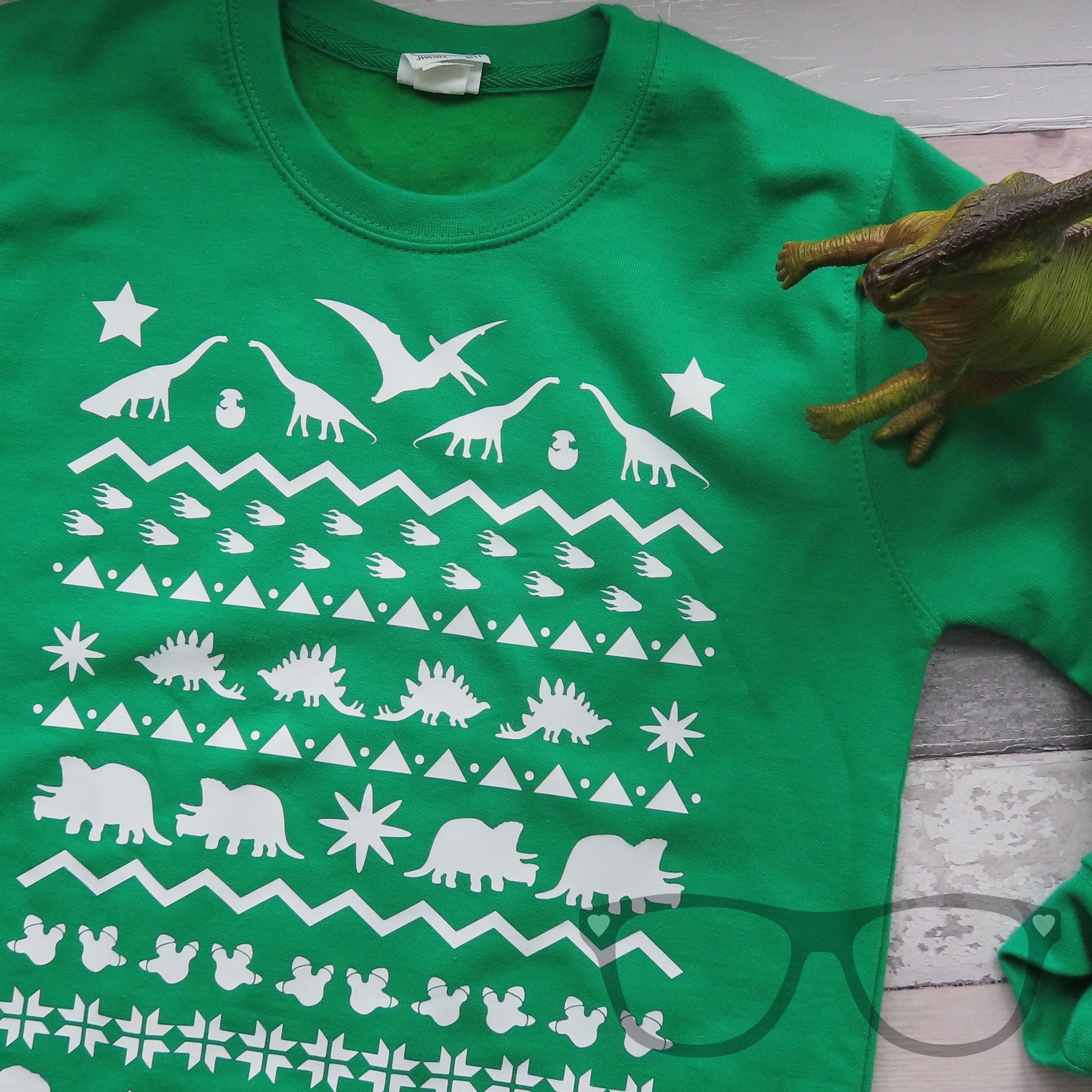 A close up of a green sweater with white dinosaurs heat pressed onto the front. Irene the Parasaurolophus is standing over the jumper.