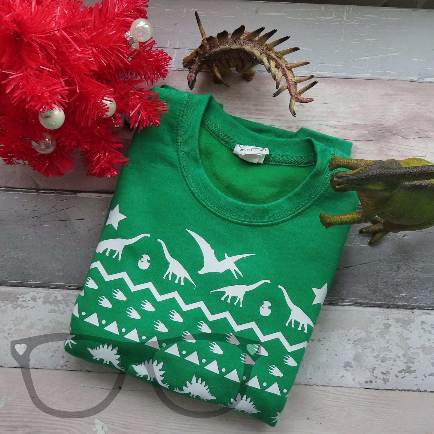 A green Christmas sweater with a white vinyl design on it. It is folded and shown with a Christmas tree and some dinosaurs