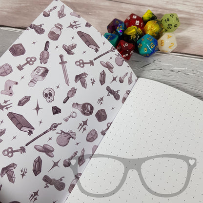 An open A6 notebook showcasing dot grid pages, with the inside cover featuring a black and white version of the front cover's pattern, depicting items for a DnD adventure.