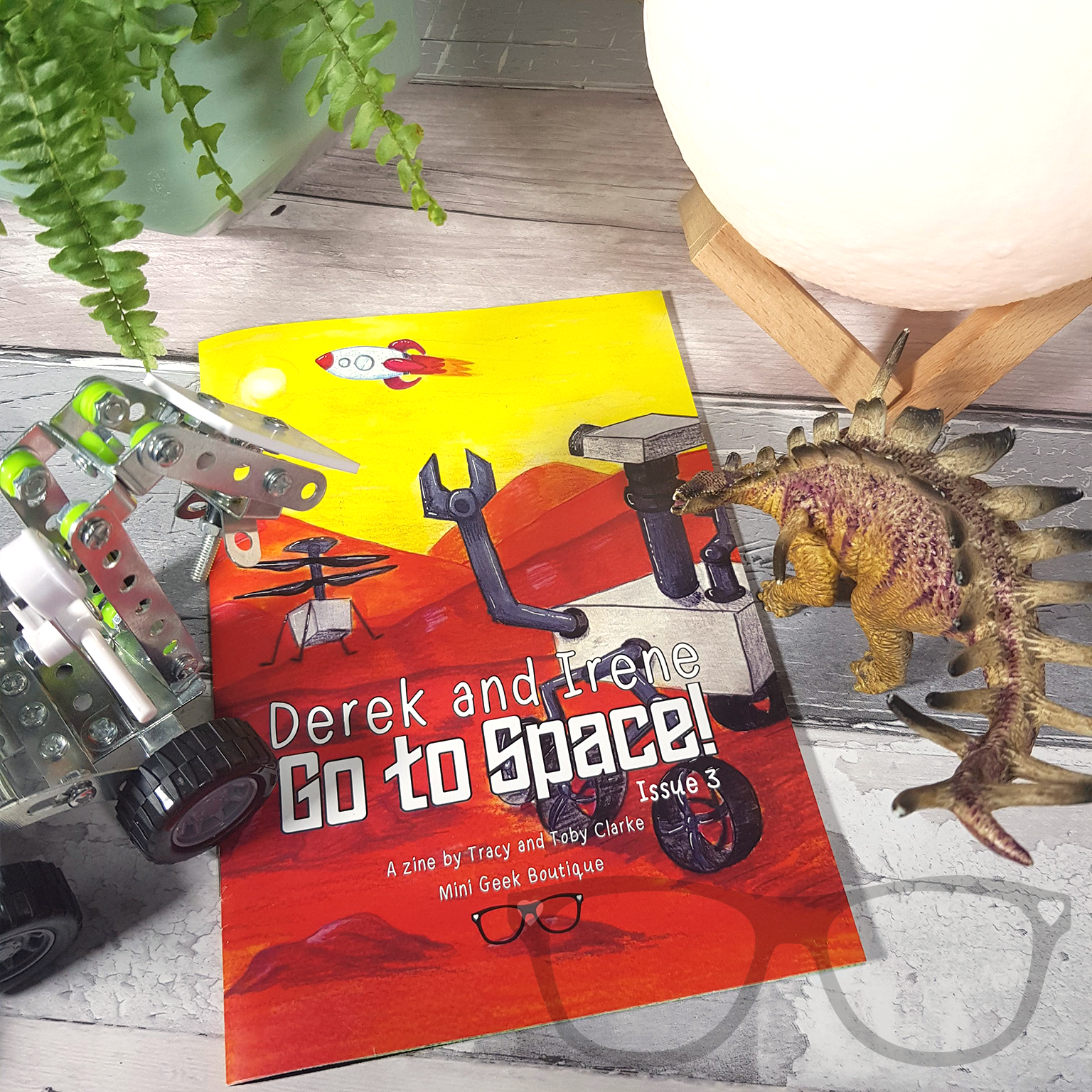 Issue 3 of the Derek and Irene zine placed alongside a Mars rover model and moonlight prop, showcasing the space adventure theme of the zine.