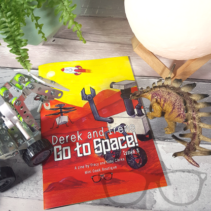 Issue 3 of the Derek and Irene zine placed alongside a Mars rover model and moonlight prop, showcasing the space adventure theme of the zine.
