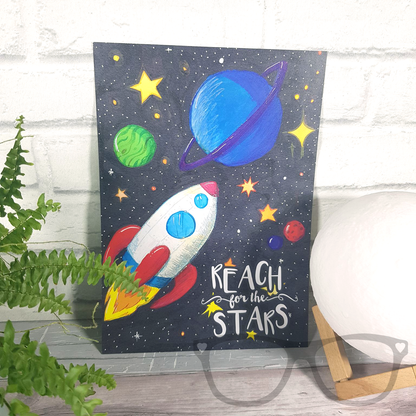 Reach for the stars A4 art print, a digital print of an original illustration showing a white rocket shooting through space with p[lanets and stars. The text reads "Reach for the stars". It is shown unframed leaning up against a white brick wall with a plant and moon lamp.