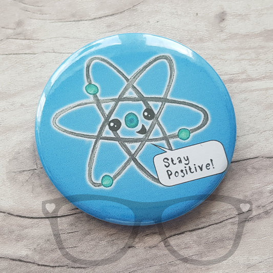 Blue 58mm circular badge with a hand drawn Atom with a face saying "Stay Positive" by Mini Geek Boutique